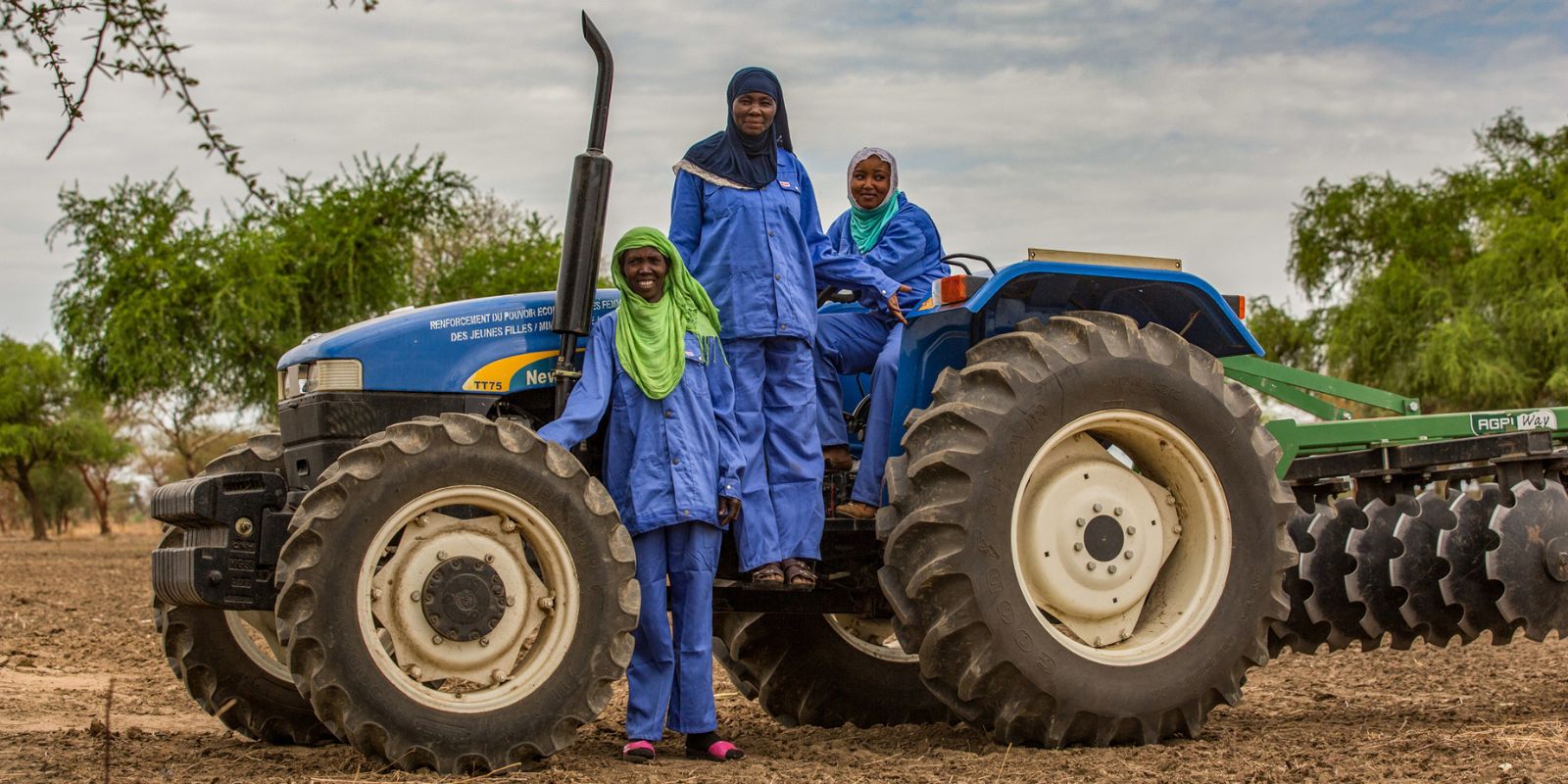 Three people in blue coveralls and green headscarves operating a New Holland tractor in a rural agricultural field, with trees and a cloudy sky in the background — representing women's active role in farming.