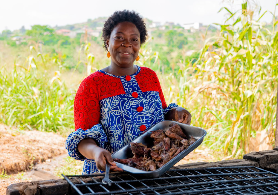 A woman wearing a colorful patterned outfit holds a metal tray filled with cooked meat pieces over an outdoor grill. The setting is an open area with tall grass and plants in the background, and distant buildings visible on a hillside.